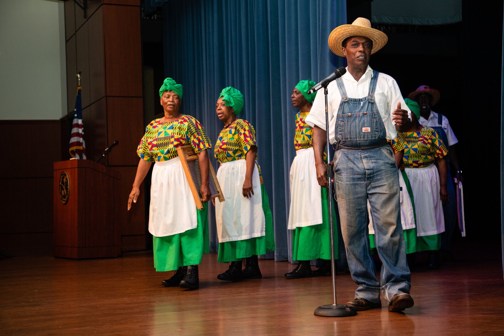 Performers in vibrant African print clothing with green headwraps and white aprons standing together on stage during a cultural presentation
